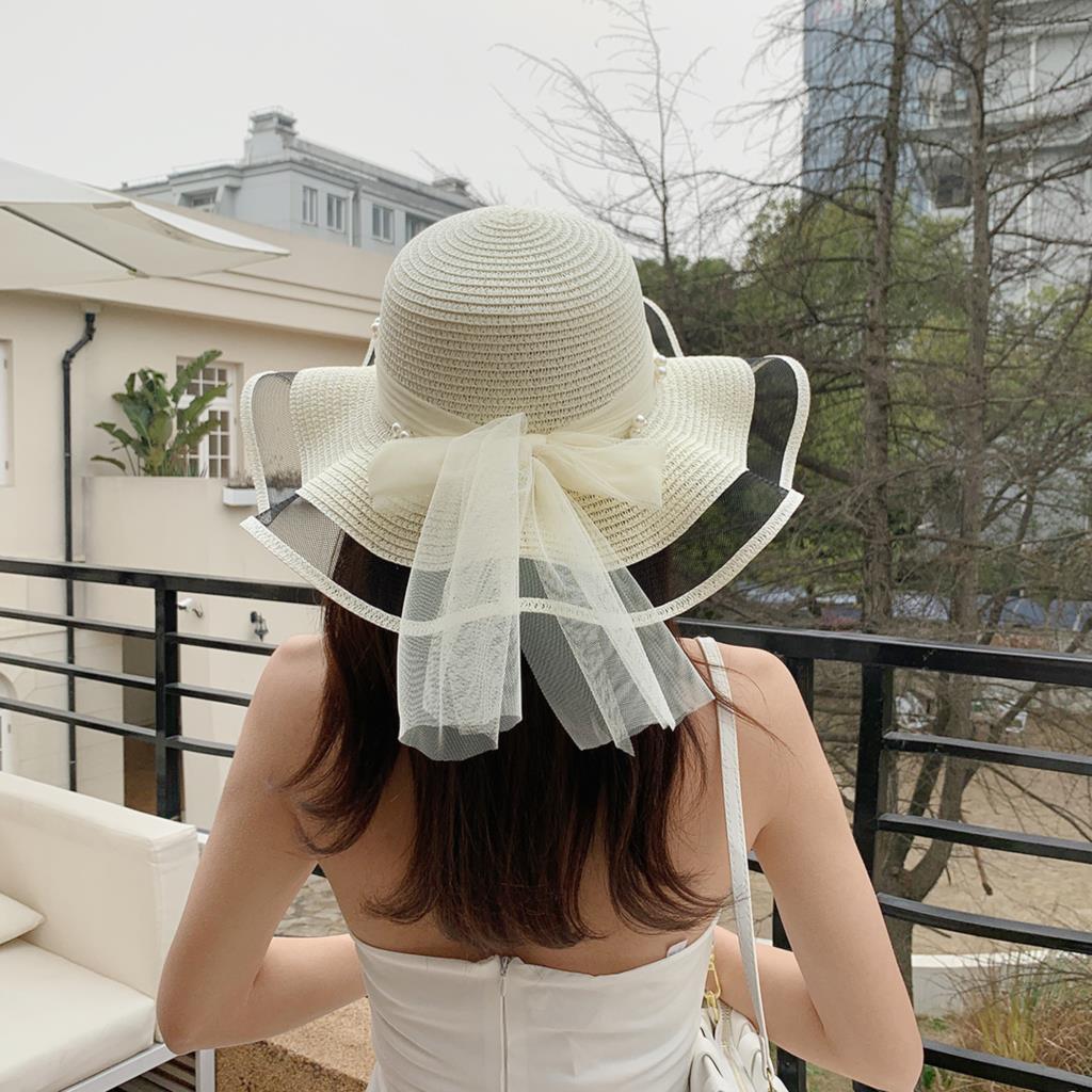 A woman wearing a white Beachy Cover Ups Big Brim Straw Beach Hat With Bow and Pearls on a balcony.