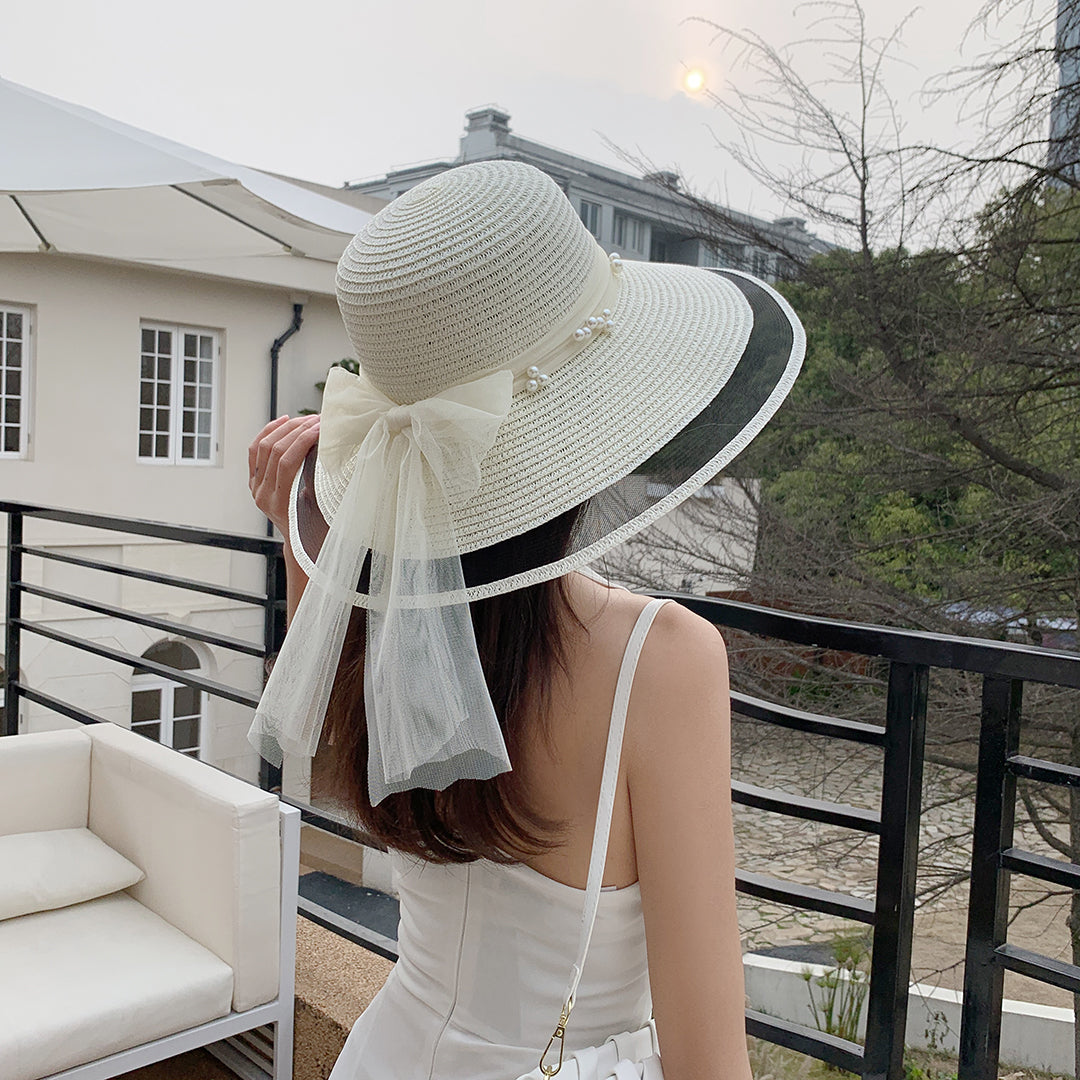 A woman wearing a Big Brim Straw Beach Hat With Bow and Pearls from Beachy Cover Ups on a balcony.