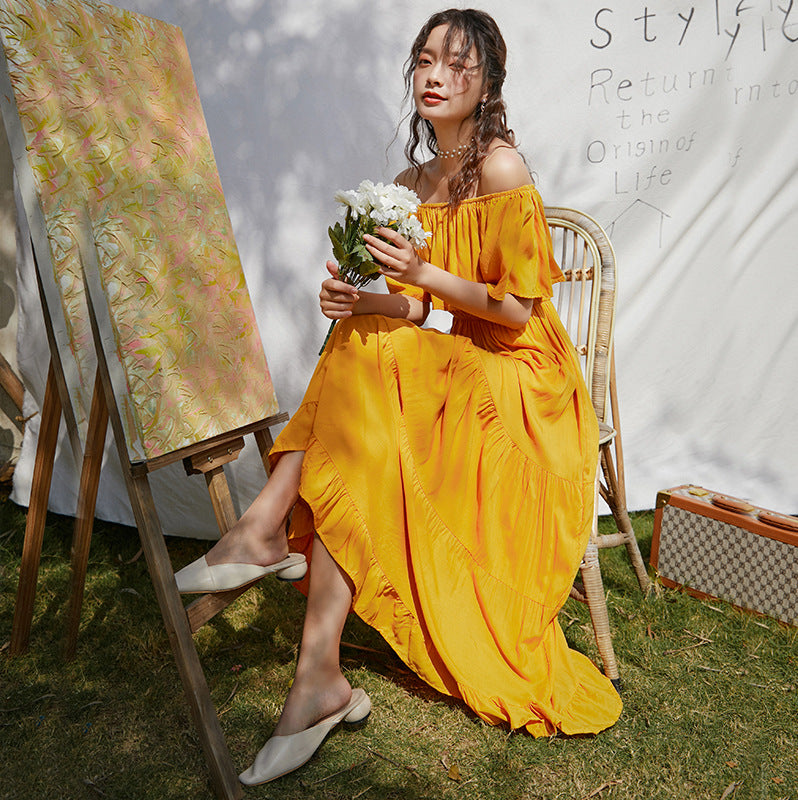 A woman in a yellow Beachy Cover Ups one-shoulder beach umbrella dress sitting next to an easel.